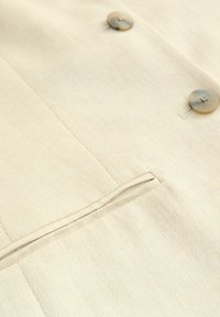 Helles beiges Gewebe mit feiner gerippter Textur, ausgestattet mit zwei runden Knöpfen und einem aufgesetzten Taschen-Detail auf der Vorderseite.