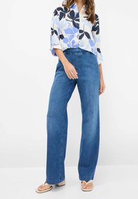 Camisa de botón de gran tamaño en azul claro con estampado floral combinada con pantalones vaqueros de pierna ancha en azul y sandalias planas color beige, enfatizando un ajuste relajado.