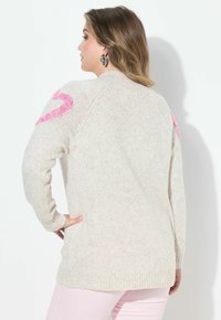 Frau mit hellbraunen Haaren, die einen beige gestrickten Pullover mit pinken Details an den Ärmeln und hellrosa Hosen trägt, steht vor weißem Hintergrund.