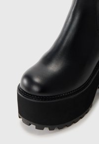 Fekete bőr platform csizma lekerekített orral, texturált felülettel, és kiemelkedő futófelülettel a vastag gumitalpon. Varrott részletek a szélén.