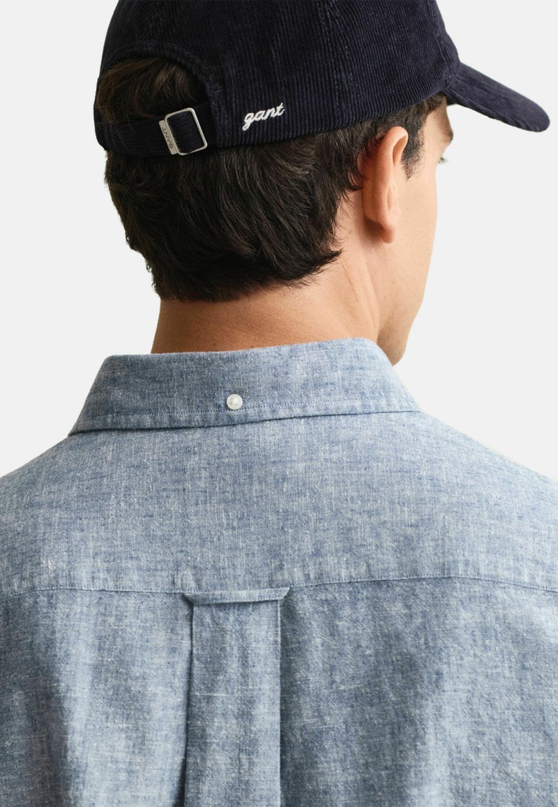 Camicia button-up azzurro chiaro, texturizzata, con pieghe posteriori e colletto, abbinata a un cappello in velluto a coste scuro. Il cappello presenta un logo del marchio sul retro.