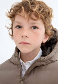 Bežna puhovka s kapuco, z zadrgo in gladko teksturo, nosi čez belo srajco s kragnico. Otrok s kodrastimi lasmi in pegicami.