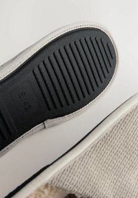 Schuhsohle mit einem glatten schwarzen Gummiunterteil, das horizontale Rillen aufweist, umrandet von einem hellfarbigen Leinensaum und einem strukturierten oberen Abschnitt.