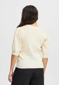 Kremkrāsas ribota džemperis ar īsām pūkainām piedurknēm, apaļu kakla izgriezumu un pieguļošu siluetu, apvienots ar melniem biksēm. Mīksta tekstūra un smalka dizaina izvēle.