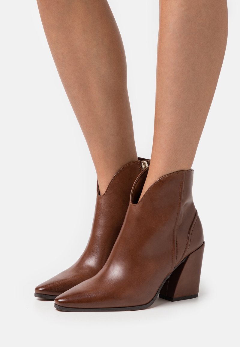 Bottes de cheville en cuir marron avec un bout pointu, un design découpé au niveau de la cheville et un talon épais. Texture lisse et détails de couture minimaux.