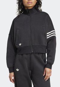 Donna che indossa una giacca nera Adidas con zip e strisce bianche su una manica, abbinata a pantaloni neri con il logo Adidas.