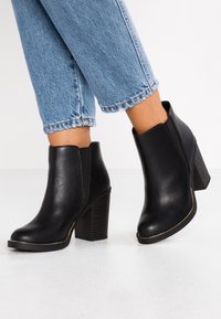 Botas de tobillo de cuero negro con un tacón alto y grueso y paneles elásticos laterales, con una puntera redondeada y un sutil diseño dorado en la suela.