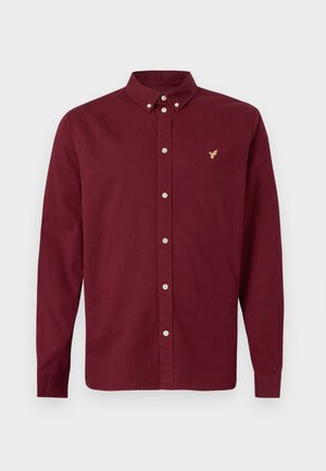 Pikkade varrukatega nööpidega pluus sügavbordoopunas, nööbitava kraega ja vasakul rinnal väikese tikitud kuldse linnu logoga.