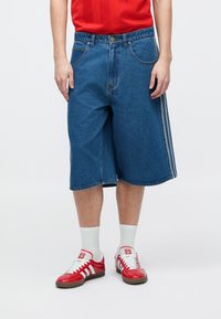 Άτομο που φοράει μπλε denim μακριά σορτς, λευκές κάλτσες, κόκκινα και λευκά αθλητικά παπούτσια Adidas και κόκκινο πουκάμισο, στέκεται μπροστά σε απλό φόντο.