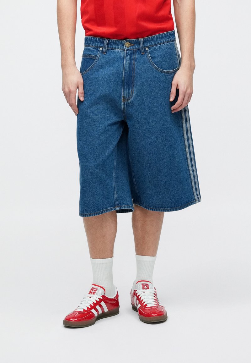 Personne portant un short long en denim bleu, des chaussettes blanches, des baskets Adidas rouges et blanches, et une chemise rouge, debout devant un fond uni.