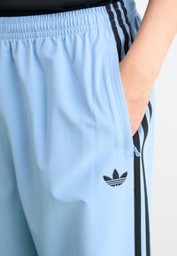 Calças desportivas azul claro feitas de um tecido suave, com riscas pretas nas laterais e um logo preto bordado na perna.