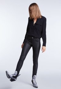Femme portant une chemise noire boutonnée, un pantalon en cuir noir et des bottines en cuir serpent blanc et noir, levant une jambe sur un fond uni.