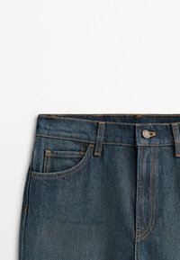 Jean en denim bleu foncé avec une coupe droite, présentée avec des coutures en cuivre, une poche avant unique et une fermeture à bouton à la taille.