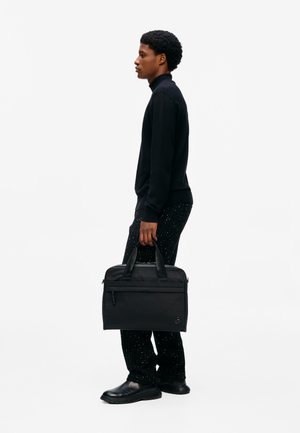 Hombre que lleva un suéter negro y pantalones negros con estampado sostiene un maletín negro al lado, de pie contra un fondo blanco liso.