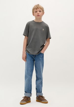 Graues Baumwoll-T-Shirt mit kurzen Ärmeln und einem kleinen Emblem, kombiniert mit hellblauen Denim-Jeans und braunen Sneakers mit hellbraunen Akzenten.
