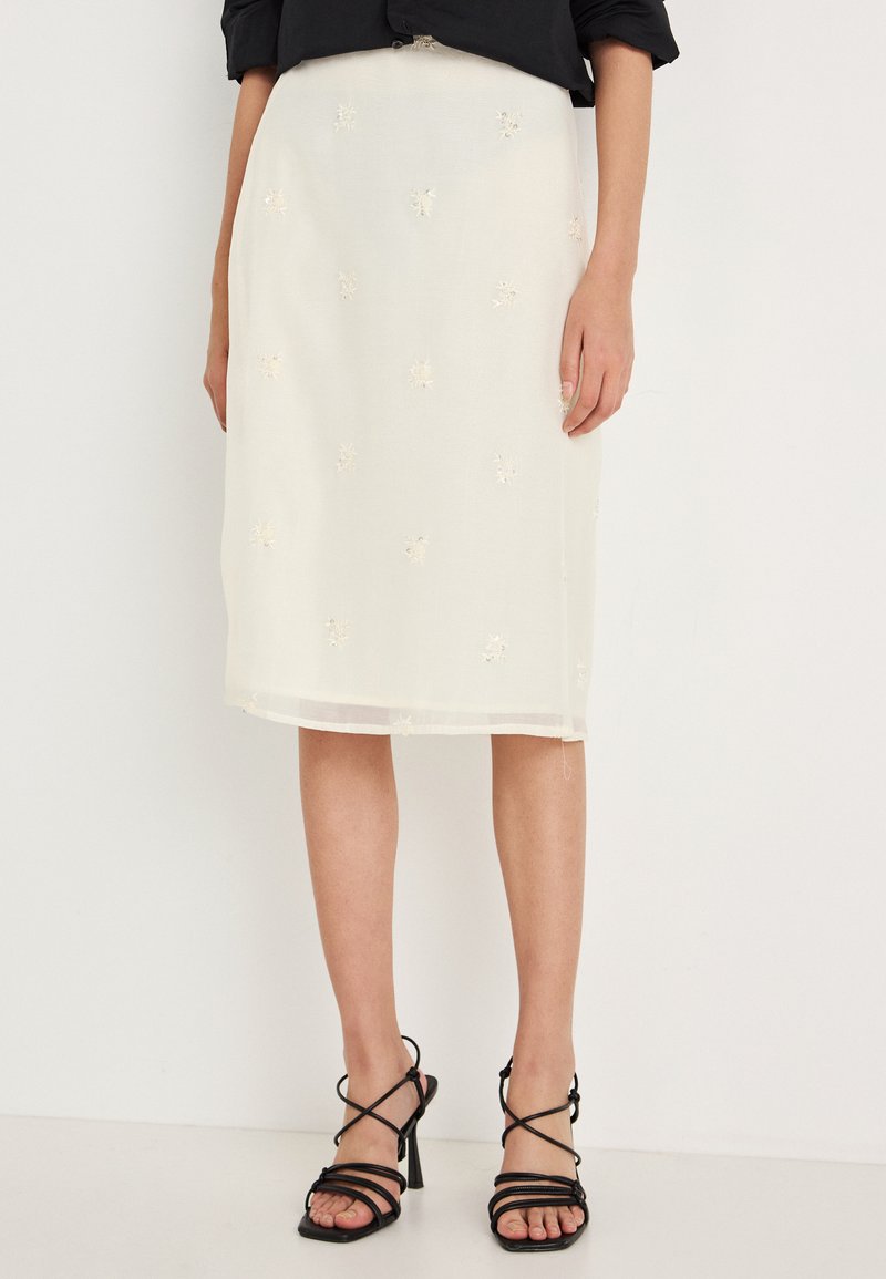 Saia midi branca com padrões florais bordados. Feita de um tecido leve, apresenta um corte reto e uma bainha transparente. Combinada com sandálias pretas de tiras.