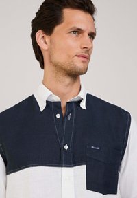 Camisa de cuadros azul y blanca, mitad lino azul marino y mitad tela blanca, con un bolsillo en el pecho y cierre de botones con costuras en contraste.