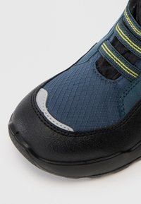 Nærbillede af en blå og sort sneakersnude med struktureret mesh-stof, sort gummisål og blå snørebånd med gule striber.