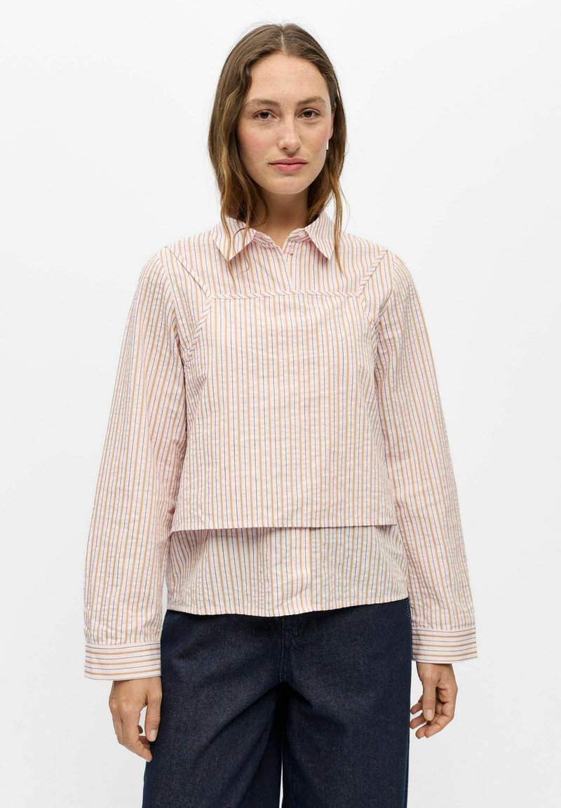 Femme à la peau claire et aux cheveux bruns, portant une chemise à manches longues rayée claire et un pantalon foncé, debout devant un fond blanc uni.