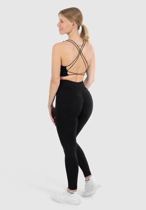 Vrouw die staat, glimlachend opzij, gekleed in een zwarte sportbeha met bandjes, hoge zwarte leggings en witte sport schoenen op een grijze achtergrond.
