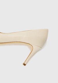 Beige high-heeled shoe with a smooth leather finish, tapered toe, and metallic gold-accented heel. No visible patterns or embellishments.