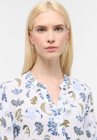 Blusa bianca con motivi floreali blu e verdi. Presenta un collo a V e un tessuto leggero e uniforme. Design pulito e minimalista.