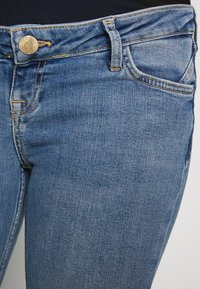 Jeans em denim azul-escuro com um corte slim, apresentando um botão de latão, costura visível e bolsos frontais. Textura suave e design clássico.