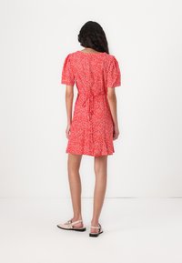 Rode jurk met een wit bloemenpatroon, korte pofmouwen, een aansluitende taille met een strik en een uitlopende rok. Draagt met beige sandalen.