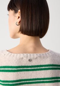 Maglione crema con strisce orizzontali verdi, scollo rotondo e texture a coste. Presenta un piccolo tag metallico rotondo sul retro dello scollo.
