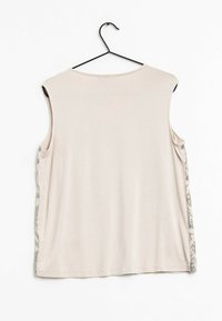 Haut beige sans manches sur un cintre noir, avec des ornements en sequins argentés courant le long des côtés, sur un fond blanc.