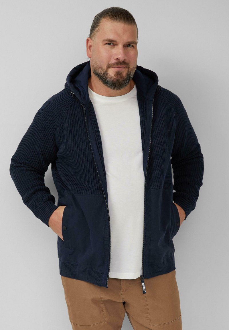 Mężczyzna z brodą noszący granatowy rozpinany sweter z dzianiny na białej koszulce i br ązowych spodniach, stojący z rękami w kieszeniach na jednolitym tle.