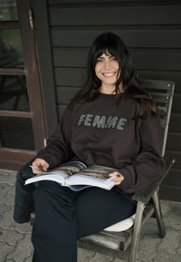 Felpa marrone con la scritta "FEMME" in rilievo, indossata con pantaloni neri. Persona seduta su una sedia di legno, che tiene in mano un libro aperto.