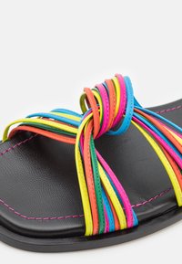 Sandale plate noire avec des lanières tressées vibrantes en rose, bleu, jaune et vert. Finition avec des coutures roses remarquables le long des bords.