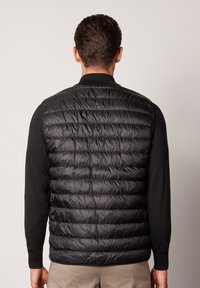 Svart pufferväst med kviltad design, utan ärmar, lätt och med ståkrage och elastisk nederkant, bärs över en svart långärmad topp.