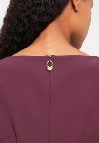 Rochie bordo cu decolteu bateau, fermoar la spate și un accent din hardware circular auriu la gât. Material texturat cu un finisaj neted.