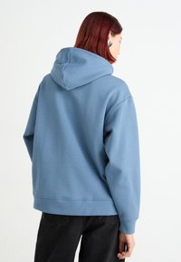 Modrá mikina s kapucí vyrobená z měkkého materiálu, s velkou kapucí a žebrovanými manžetami, spárovaná s černými džíny. Design je jednoduchý a uvolněný.