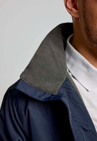 Giacca blu navy con colletto in pile grigio, caratterizzata da una texture liscia e un design del colletto tradizionale, che rivela parzialmente una camicia bianca sottostante.