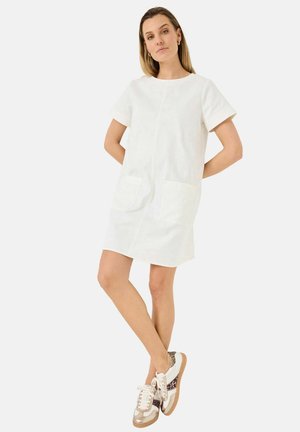 White short-sleeve dress with round neckline, two front pockets, and a straight cut. Paired with beige and patterned sneakers.