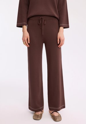 Osoba nosząca brązowe dzianinowe spodnie o szerokich nogawkach z troczkiem w pasie, dopasowany top, złoty pierścionek oraz złote plecione płaskie buty z czarnym elastycznym paskiem.