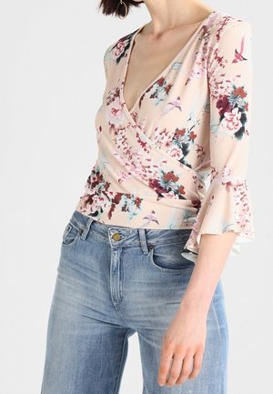 Kobieta ubrana w jasnoróżową kwiecistą bluzkę wiązaną z rozszerzanymi rękawami, zestawioną z niebieskimi jeansami z wysokim stanem i złotymi kolczykami kołami.