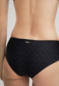 Schwarzer Bikini-Höschen mit einem subtilen Muster und glatter Textur. Enthält ein kleines goldfarbenes Logo-Tag auf der Rückseite.