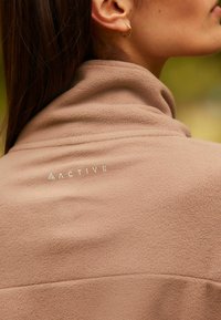 Bruine fleece jas met een hoge kraag. Beschikt over een gestructureerde stof en een logo op de achterkant. Eenvoudig ontwerp, getailleerde pasvorm.