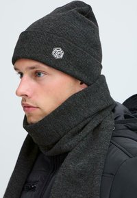 Nuori mies, joka käyttää tummanharmaata neulottua myssyä ja siihen sopivaa huivia mustan takin kanssa, kääntyy vasemmalle yksinkertaista vaaleaa taustaa vasten.