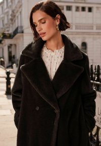 Palton negru din lână, cu două rânduri de nasturi și guler lat, cu o textură moale și două capse. Purtați peste o bluză albă cu volane și accente aurii.