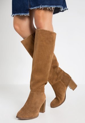 Jambes portant des bottes en daim beige montant jusqu'aux genoux avec des talons blocs empilés et une jupe en jean à ourlet effiloché sur un fond uni.