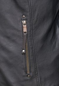 Detail einer schwarzen Lederjacke mit einer Reißverschlusstasche, messingfarbenem Reißverschluss, braunem Leder-Ziehebändchen und silbernem Druckknopf-Akzent. Glatte Textur.
