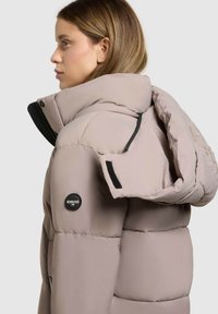 Steppjacke in hellem Beige mit sichtbarer Steppstruktur, schwarzen Beschlägen und abnehmbarer Kapuze, mit einem Logo am Ärmel.