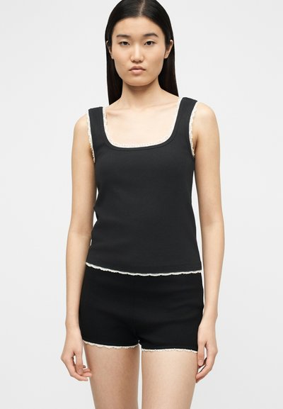 Jeune femme portant un haut noir sans manches et un short assorti avec une bordure en dentelle blanche, se tenant devant un fond clair uni.