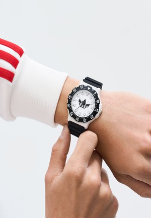 Črno-bela športna ura s krožnim številčnikom, črnim silikonskim trakom in rotirajočim obročem. Roka nastavlja čas. Bela ozadje.