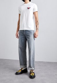 White cotton t-shirt with red and black graphic on the chest, paired with light blue denim jeans and black loafers featuring floral embroidery.